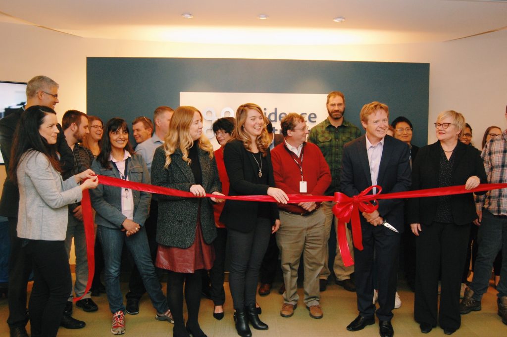 A group of people standing in front of the Evidence Partners logo displayed at their newly expanded office in Kanata, Ontario. The team prepares to cut the ribbon.