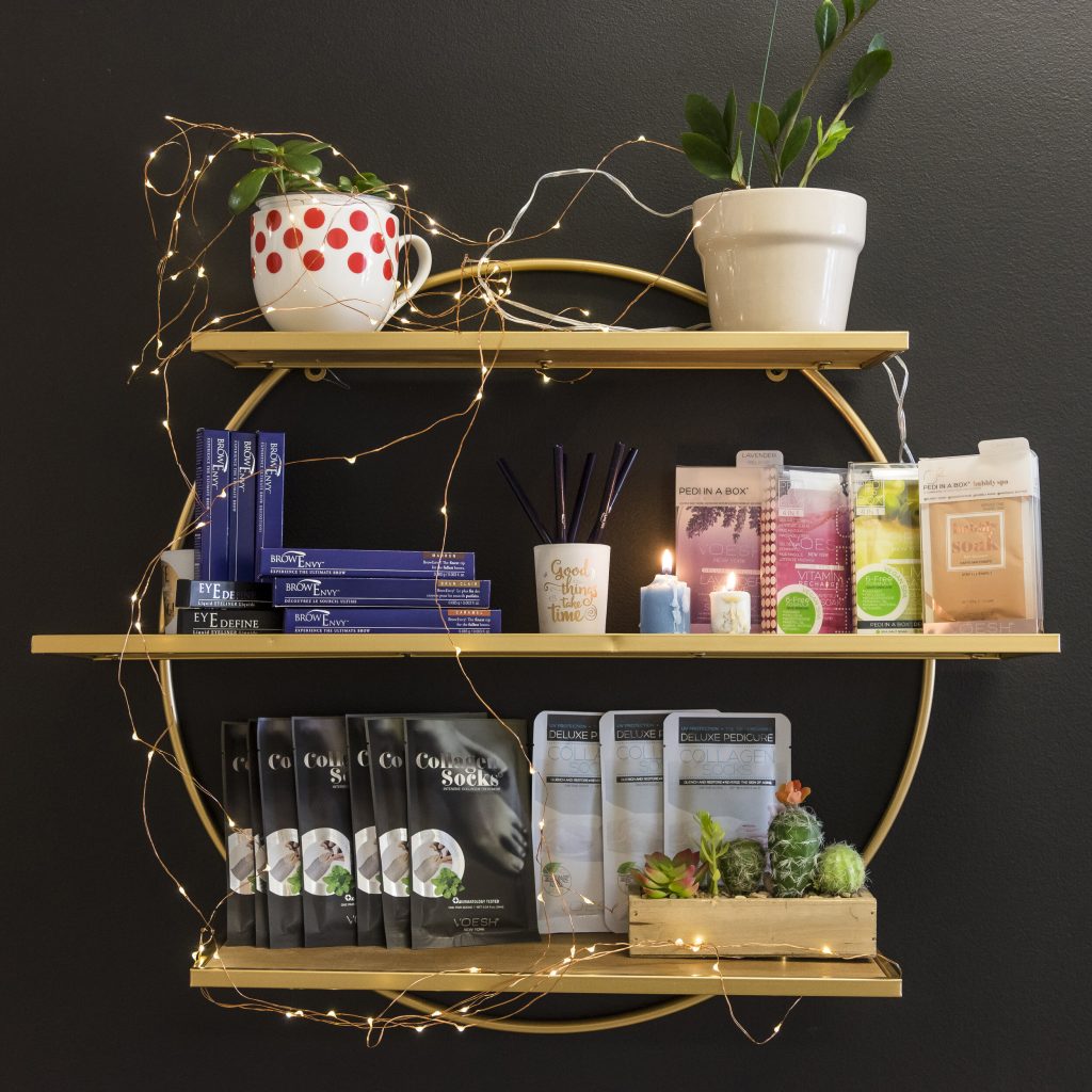 An assortment of items found in the Esthetics By Lucie shop, arranged on three shelves and wrapped in decorative wire lighting. 