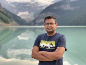 Developer Kazi Ahmed stands with his arms crossed in front of a light blue lake amidst misty mountains. 
