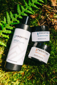 Three containers of Frontier Skincare products are arranged in grass and leaves. 