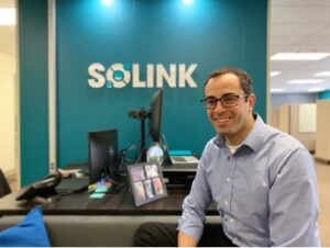 Solink CEO Mike Matta leans against a chair in front of blue wall featuring the company logo. 